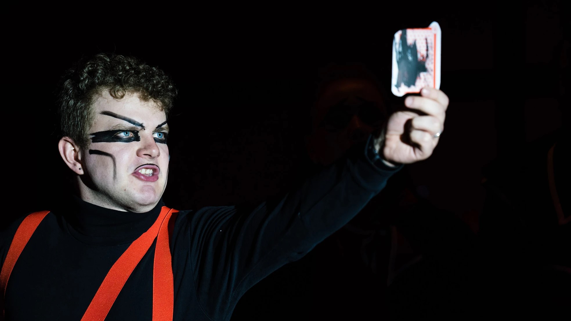 A dramatic photo of Chris Murphy with black features painted on and wearing red braces in Cumbria Opera Group's production of Barber's a Hand of Bridge